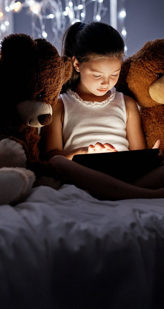 Kind liest auf einem Tablet im Bett, umgeben von Teddybären, Ausdruck von Ökostromnutzung bei Nacht.