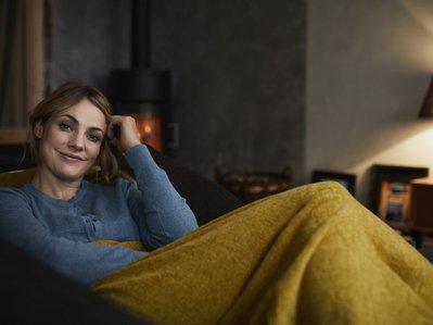 Eine Frau sitzt entspannt auf dem Sofa unter einer gelber Decke. Eine Tischlampe steht für den Stromverbrauch von Licht.