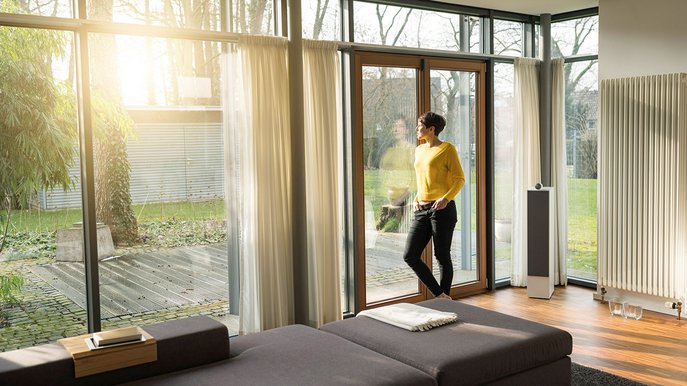 Gasverbrauch Einfamilienhaus: Frau steht am Fenster, Sonnenlicht, moderner Wohnraum, große Heizung, Energieeffizienz im Fokus.