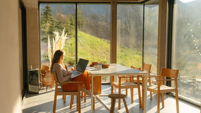 Frau arbeitet am Laptop in einem sonnendurchfluteten Raum mit Panoramablick in die Natur. Symbolisiert Informationen zum Glasfaser-Wechselservice von ENTEGA.