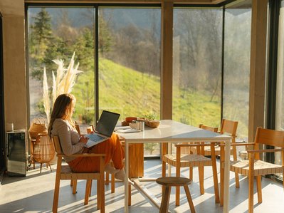 Frau arbeitet am Laptop in einem sonnendurchfluteten Raum mit Panoramablick in die Natur. Symbolisiert Informationen zum Glasfaser-Wechselservice von ENTEGA.