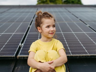 Ein Mädchen in gelbem Kleid sitzt vor Solarmodulen. Sie stehen als Symbol für die Zukunft der Energie und den Kohleausstieg.