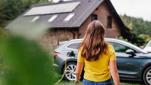 Frau in gelbem Pullover nähert sich einem Elektroauto, das vor einem Haus mit Solarpanelen steht. Kontext: Smartes Laden mit dynamischem Stromtarif.