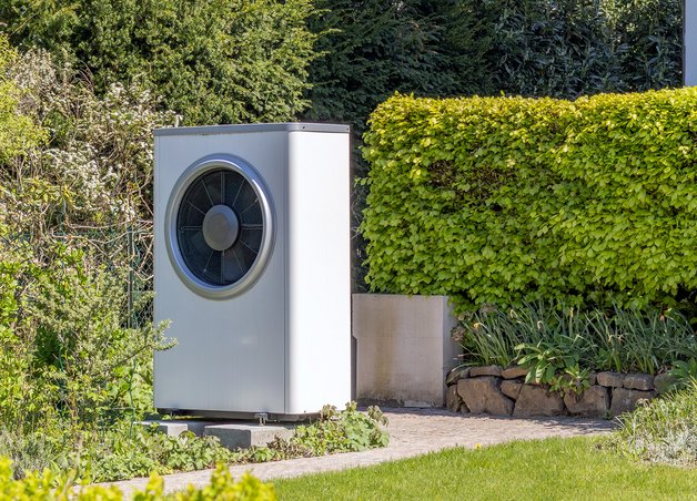 Moderne Wärmepumpe steht frei im Garten, umgeben von Hecken und Pflanzen. Strom Wärmepumpe relevant für Energieeffizienz.