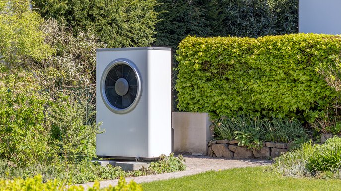 Moderne Wärmepumpe steht frei im Garten, umgeben von Hecken und Pflanzen. Strom Wärmepumpe relevant für Energieeffizienz.