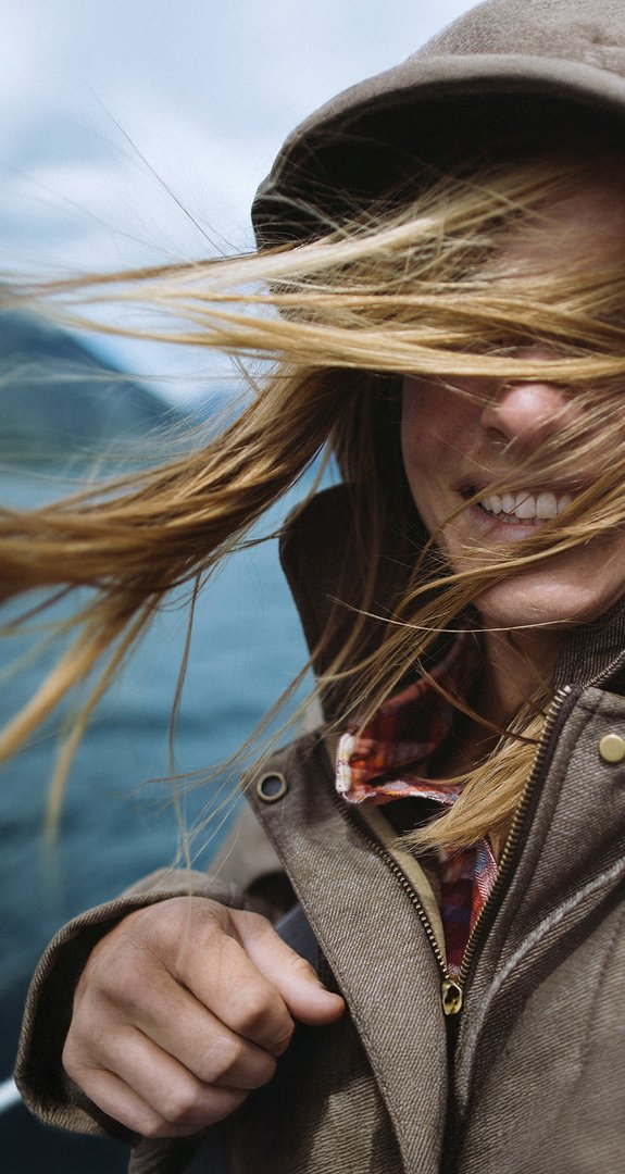 Person mit Kapuze, vom Wind verwehte Haare, lächelnd. Wasserlandschaft deutet Verbindung zur Natur durch 100 % Ökostrom von ENTEGA an.
