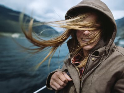 Person mit Kapuze, vom Wind verwehte Haare, lächelnd. Wasserlandschaft deutet Verbindung zur Natur durch 100 % Ökostrom von ENTEGA an.