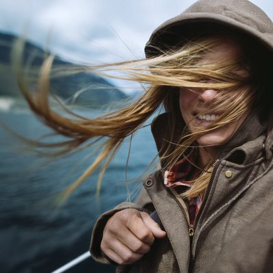Person mit Kapuze, vom Wind verwehte Haare, lächelnd. Wasserlandschaft deutet Verbindung zur Natur durch 100 % Ökostrom von ENTEGA an.