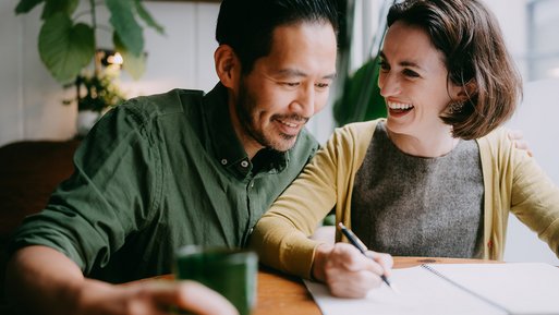 Lächelndes Paar am Tisch, plant mit Notizbuch. Darstellung von finanzieller Förderung für nachhaltige Energieoptionen.