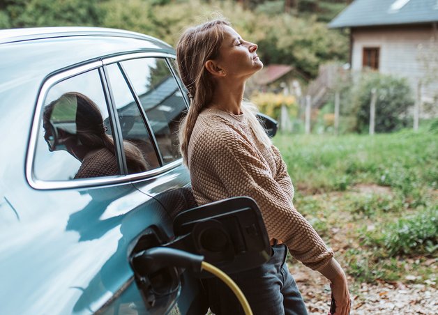 Frau lehnt entspannt am Elektroauto, das umweltfreundlich zu Hause geladen wird; grüne Umgebung betont Elektroauto Umwelt.