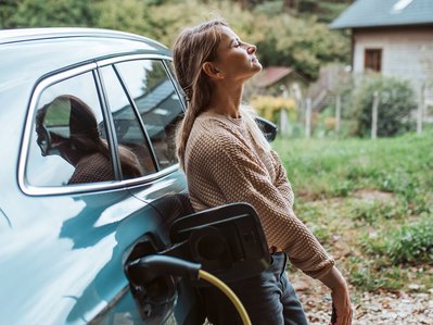 Frau lehnt entspannt am Elektroauto, das umweltfreundlich zu Hause geladen wird; grüne Umgebung betont Elektroauto Umwelt.