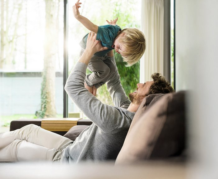 Mann hebt lachendes Kind hoch, helle Wohnzimmerumgebung. Darstellung von Wärme und Freude durch effizientes Heizen mit Wärmepumpe.