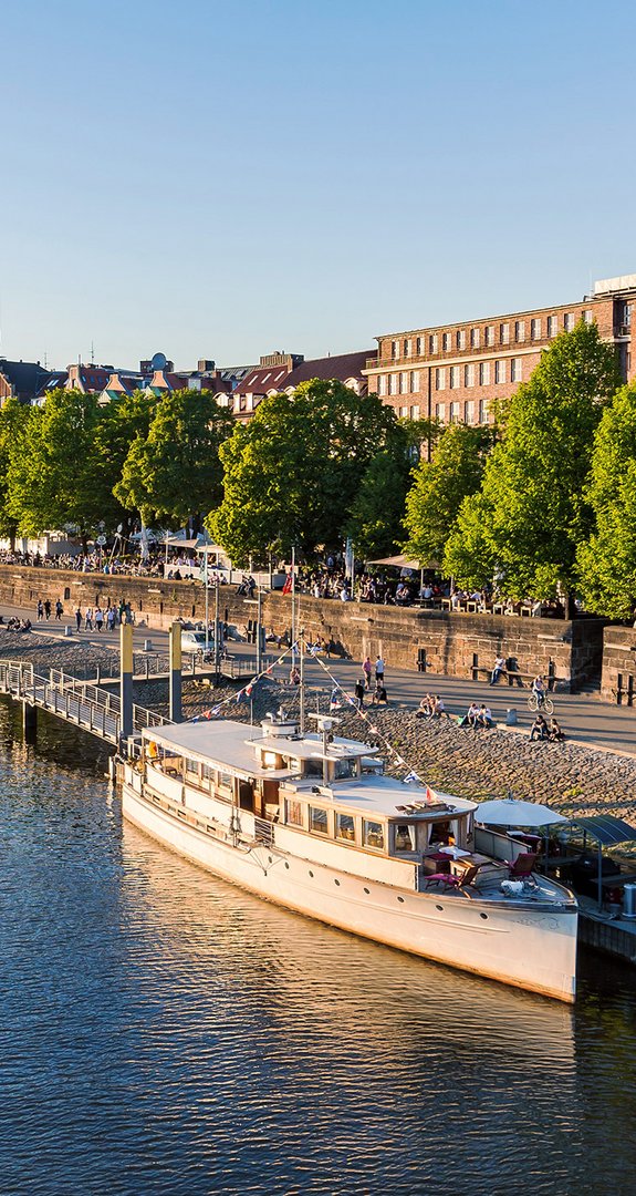Lebendige Bremer Weserpromenade mit Schiffen, Ruderboot und grünen Bäumen, steht für Lebensqualität und Ökostrom Bremen im urbanen Raum.