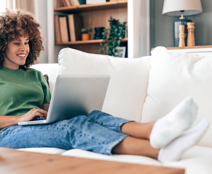 Frau sitzt auf Sofa und nutzt Laptop, symbolisiert Nutzung der digitalen Assistentin Ella.