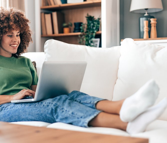 Frau sitzt auf Sofa und nutzt Laptop, symbolisiert Nutzung der digitalen Assistentin Ella.