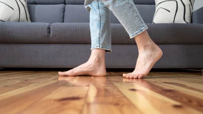 Füße laufen barfuß über warmen Holzboden vor Sofa, Stromverbrauch Fußbodenheizung als zentrales Thema für Wohnkomfort.