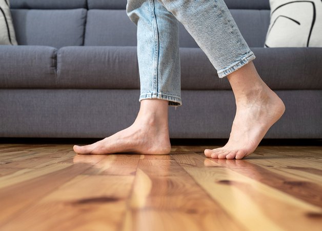 Füße laufen barfuß über warmen Holzboden vor Sofa, Stromverbrauch Fußbodenheizung als zentrales Thema für Wohnkomfort.