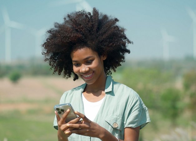 Frau mit Smartphone vor Windkraftanlagen in sonniger Landschaft; Symbol für erneuerbare Energieversorgung.