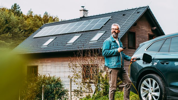 Bild zeigt ein Einfamilienhaus mit Solaranlage und davor einen Mann, der sein E-Auto auflädt