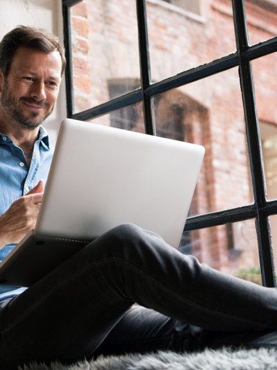 Bild zeigt einen Mann, der mit Laptop auf der Fensterbank sitzt – Thema: Stromrechnung.