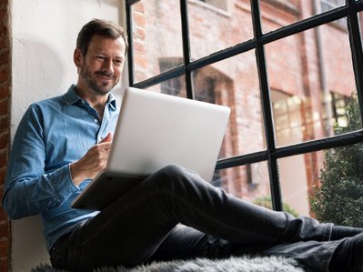 Bild zeigt einen Mann, der mit Laptop auf der Fensterbank sitzt – Thema: Stromrechnung.