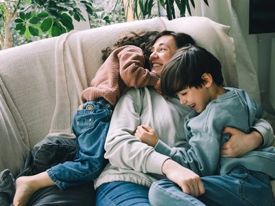 Lächelnde Mutter kuschelt mit zwei Kindern auf einem Sofa, umgeben von Pflanzen. Ausdruck von Liebe und familiärer Geborgenheit in entspannter Atmosphäre.