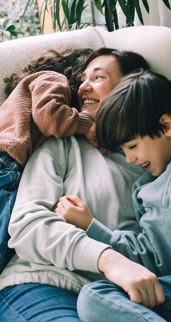 Lächelnde Mutter kuschelt mit zwei Kindern auf einem Sofa, umgeben von Pflanzen. Ausdruck von Liebe und familiärer Geborgenheit in entspannter Atmosphäre.