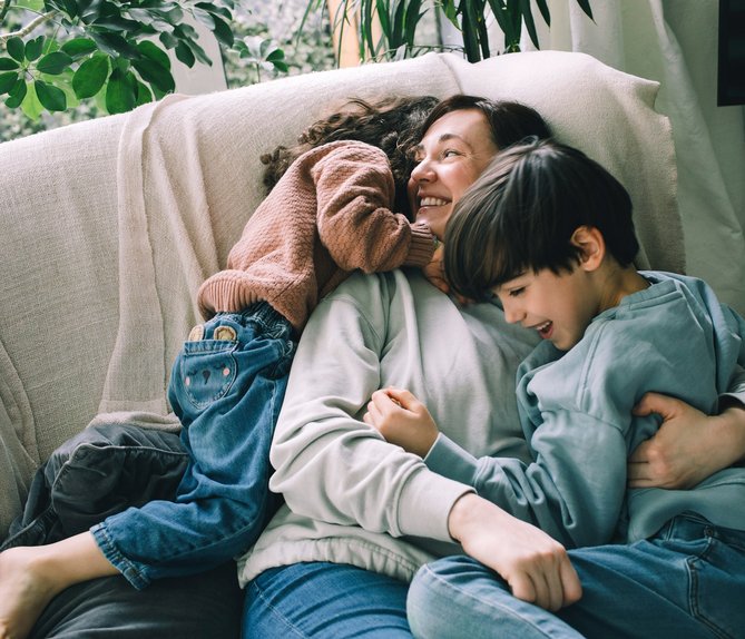 Lächelnde Mutter kuschelt mit zwei Kindern auf einem Sofa, umgeben von Pflanzen. Ausdruck von Liebe und familiärer Geborgenheit in entspannter Atmosphäre.
