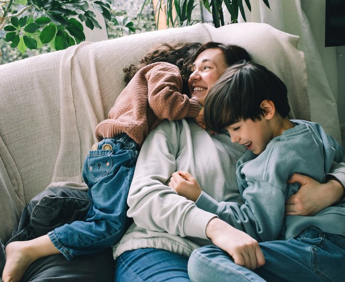 Lächelnde Mutter kuschelt mit zwei Kindern auf einem Sofa, umgeben von Pflanzen. Ausdruck von Liebe und familiärer Geborgenheit in entspannter Atmosphäre.