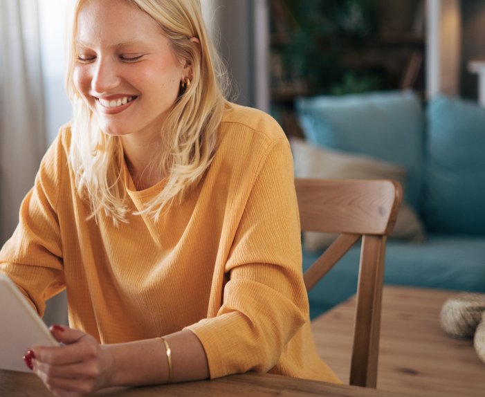 Lächelnde Frau sitzt am Tisch und benutzt ein Tablet, gemütliches Wohnzimmer. Symbolisiert einfache Suche nach Fördermöglichkeiten für Zuhause.