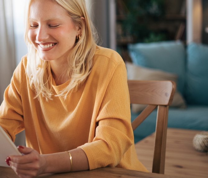 Lächelnde Frau sitzt am Tisch und benutzt ein Tablet, gemütliches Wohnzimmer. Symbolisiert einfache Suche nach Fördermöglichkeiten für Zuhause.