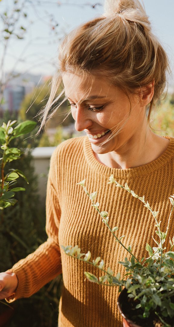 Lächelnde Person in gelbem Pullover, genießt die Sonne auf einem Balkon mit Pflanzen.
