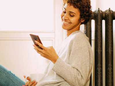 Bild zeigt eine lächelnde Frau, die auf ihr Smartphone schaut. Sie lehnt an einem Heizkörper als Symbol für die Thematik E-Heizung.