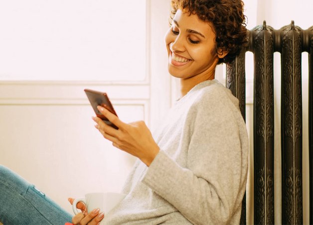 Bild zeigt eine lächelnde Frau, die auf ihr Smartphone schaut. Sie lehnt an einem Heizkörper als Symbol für die Thematik E-Heizung.
