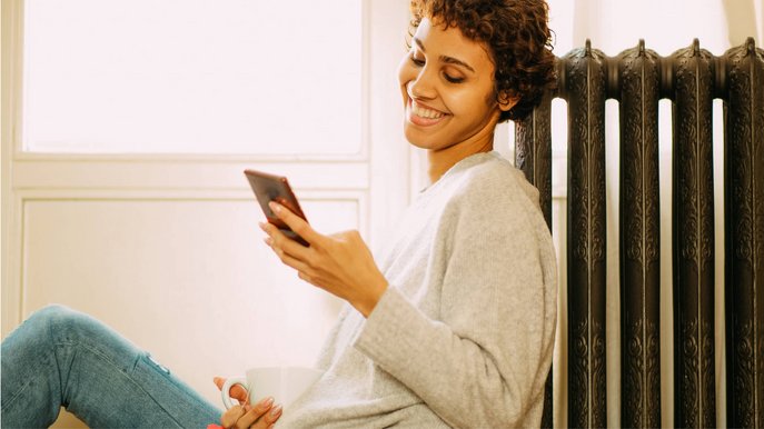 Bild zeigt eine lächelnde Frau, die auf ihr Smartphone schaut. Sie lehnt an einem Heizkörper als Symbol für die Thematik E-Heizung.