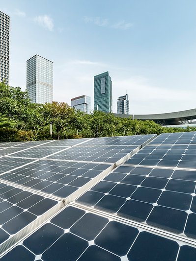 Photovoltaik-Panels in einem grünen Bereich mit modernen Hochhäusern im Hintergrund, verbindet nachhaltige Technik mit urbaner Landschaft.