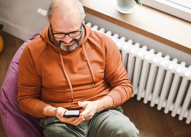 Mann sitzt mit seinem Handy vorm Heizkörper und prüft seine Heizkostenabrechnung.