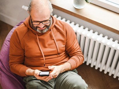 Mann sitzt mit seinem Handy vorm Heizkörper und prüft seine Heizkostenabrechnung.