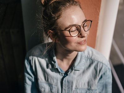 Frau mit Brille und geschlossenem Auge genießt die Sonne, lehnt an einer Wand. Ausdruck von Entspannung und Gelassenheit in einem ruhigen Moment.