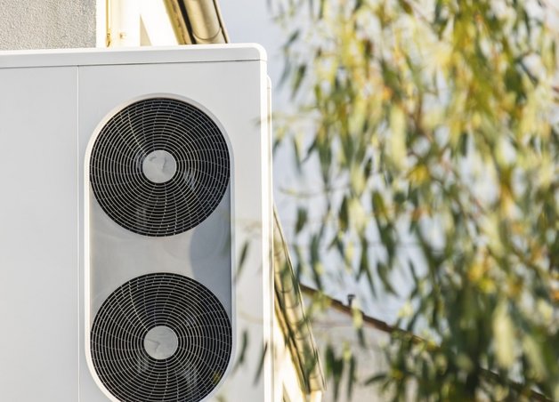 Eine Wärmepumpe, die vor einem Haus steht. Neben der Wärmepumpe sind Blätter eines Baumes zu sehen.