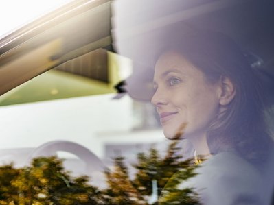 Eine Frau sitzt am Steuer hinter spiegelnder Seitenscheibe. Symbolbild für Elektroauto Nachteile wie Ladepause und Planung.