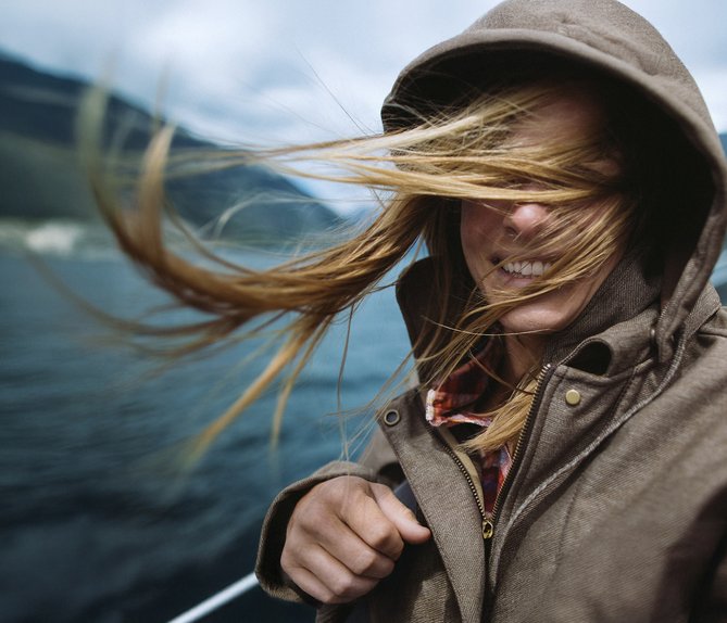 Person mit Kapuze, vom Wind verwehte Haare, lächelnd. Wasserlandschaft deutet Verbindung zur Natur durch 100 % Ökostrom von ENTEGA an.