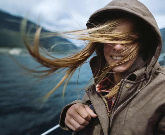 Person mit Kapuze, vom Wind verwehte Haare, lächelnd. Wasserlandschaft deutet Verbindung zur Natur durch 100 % Ökostrom von ENTEGA an.