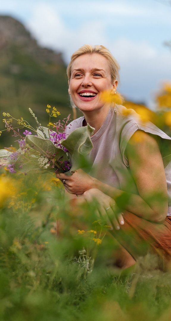 Frau lächelnd im Blumenfeld, hält bunte Blumen in der Hand, umgeben von gelben und grünen Pflanzen.