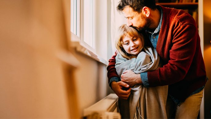 Bild zeigt einen Vater, der mit seiner Tochter vor einem winterlichen Fenster mit Heizkörper kuschelt, Symbolbild zum Thema „Gasheizung Arten“