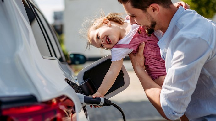 Kind steckt mit Hilfe eines Mannes das Ladekabel Elektroauto ein. Familienalltag, draußen, Freude sichtbar.