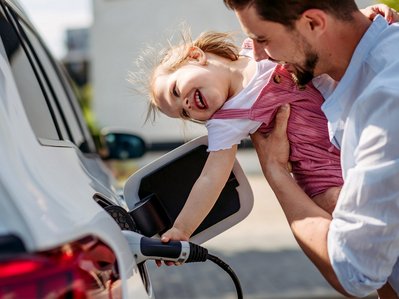 Kind steckt mit Hilfe eines Mannes das Ladekabel Elektroauto ein. Familienalltag, draußen, Freude sichtbar.