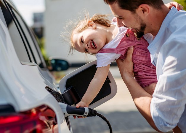 Kind steckt mit Hilfe eines Mannes das Ladekabel Elektroauto ein. Familienalltag, draußen, Freude sichtbar.