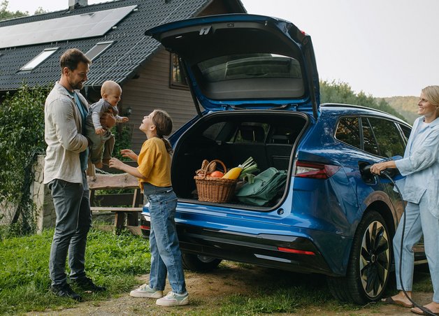 Familie lädt Elektroauto am Haus mit Solaranlage, Lebensmittel im Kofferraum; Fokus auf Elektroauto Betriebskosten.