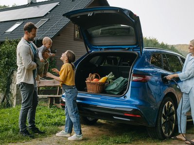 Familie lädt Elektroauto am Haus mit Solaranlage, Lebensmittel im Kofferraum; Fokus auf Elektroauto Betriebskosten.